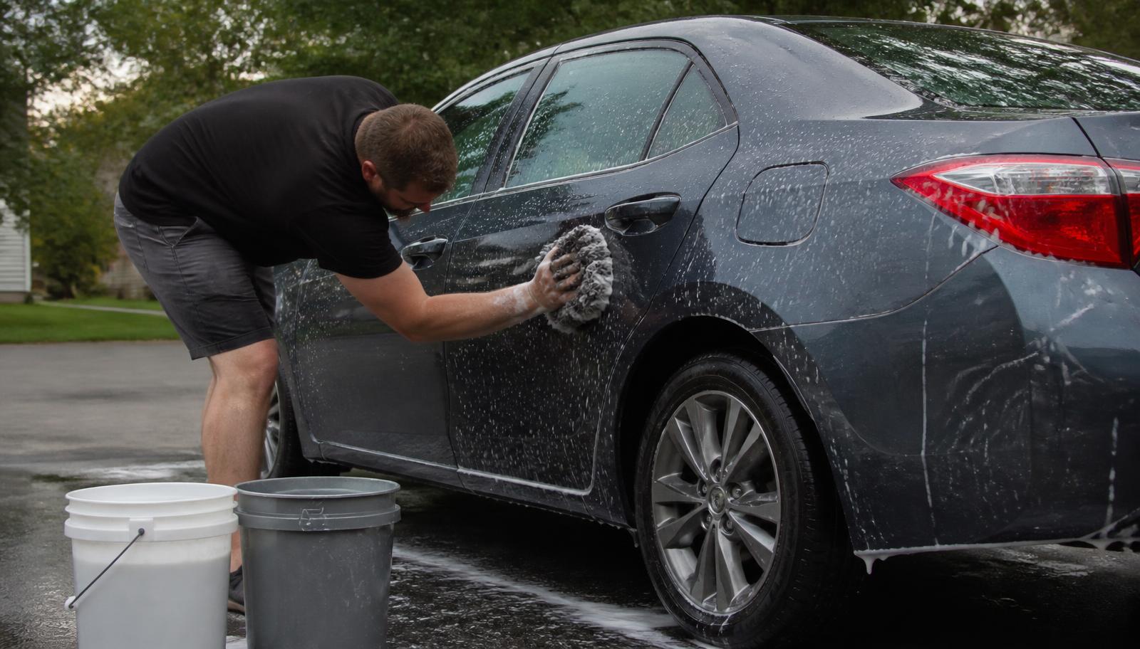 Detailer hand-washing a dark gray sedan with a wash mitt and two buckets in a Minnesota driveway