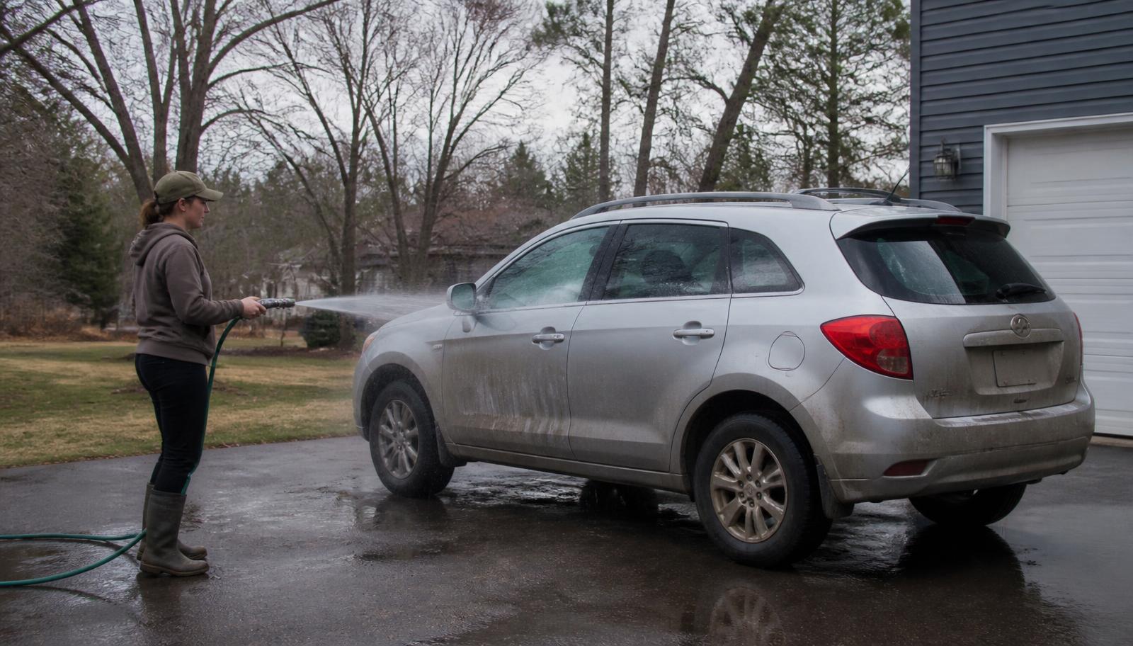 Silver crossover covered in winter road grime being rinsed in a Minnesota driveway on an early spring day